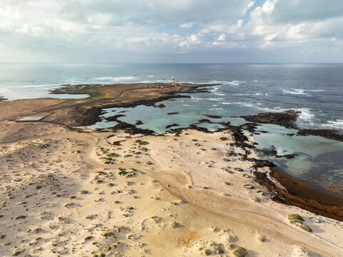Aerial view of the stark contrast between the sandy dunes and the azure waters surrounding the Toston Lighthouse, a beacon against the rugged coastline, La Oliva, Canary Islands, Spain.