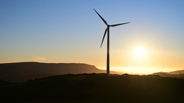 Timelapse of  wind turbine rotating at sunset over rolling hills, with the sun behind creating a dramatic backlit silhouette. Ideal for concepts of renewable energy, sustainability, landscape 