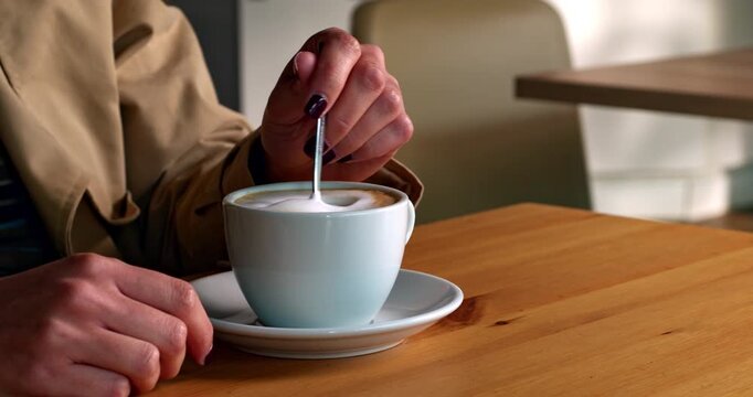 Close-up of a cappuccino being stirred in a coffee shop, as a young woman prepares to lift the cup and drink. Warm, intimate moment with soft lighting and cozy atmosphere.