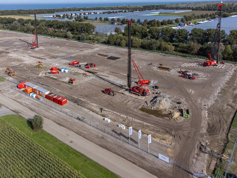Aerial view of construction site with heavy machinery contrasting against the green fields and distant blue sea, Hameiweg, Almere, Flevoland, Netherlands.