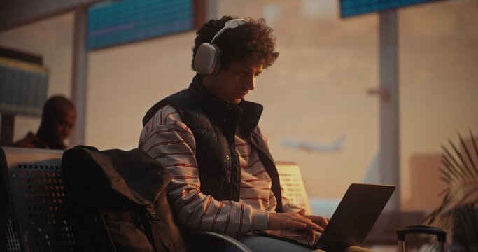 Young Man With Curly Hair Sits by Large Window in Airport Departure Lounge. Male Focused on Laptop Screen, Fingers Moving Rapidly Across Keyboard as Writes Code. Productivity of Modern Programmer.