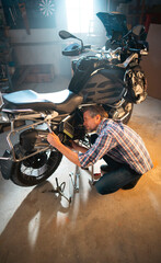 Obraz premium A man is fixing a motorcycle in his garage. He is bending down to work on the bike. Tools and accessories are on the floor. Light comes from a window.