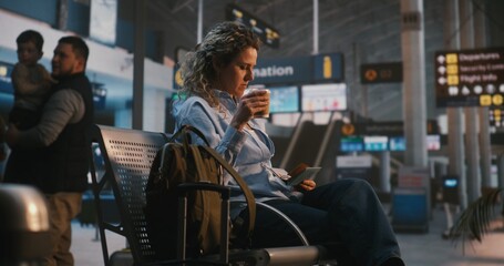 Fototapeta premium Female Traveler Arrives at Departure Hall With Backpack, Sits on Bench, and Compares Ticket With Information Board. Woman Takes Moment to Enjoy Coffee While Waiting For Boarding. Independent Travel.