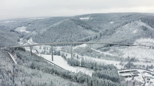 Cinematic drone shot pulling back and ascending above a long highway bridge with cars, while a train moves below through a snowy forest valley