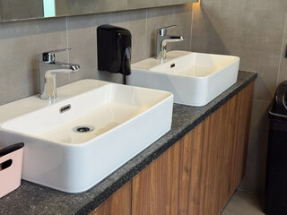 Fototapeta premium Double ceramic sinks with chrome faucets installed on stone countertop in shared restroom. Hygiene routine, simultaneous usage, public facility design and sanitation efficiency concept