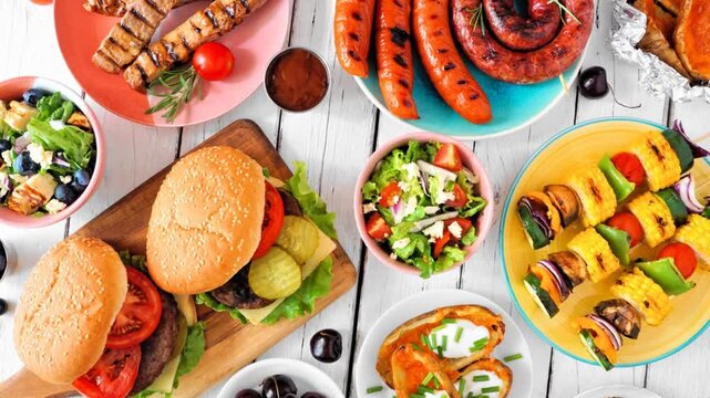 Summer BBQ or picnic food table scene. Slow rotating zoom motion. Assortment of burgers, grilled meat, vegetables, fruits, salad and potatoes. Overhead view on a white wood background.