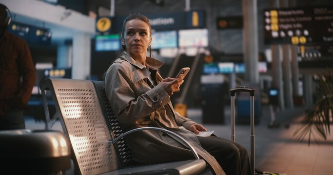 Focused Female Traveler Comparing Digital Ticket With Airport Information Board. Woman Carefully Looks Up Overhead Flight Board, Cross Referencing Gate Number and Departure Time With Data on Screen.