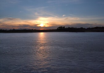 Fototapeta premium A serene sunset landscape over a partially frozen lake or wide river, with the sun reflecting on the remaining open water and ice patches.