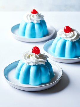 Three cool blue milk puddings in fluted molds topped with whipped cream and cherries on white plates against a light background