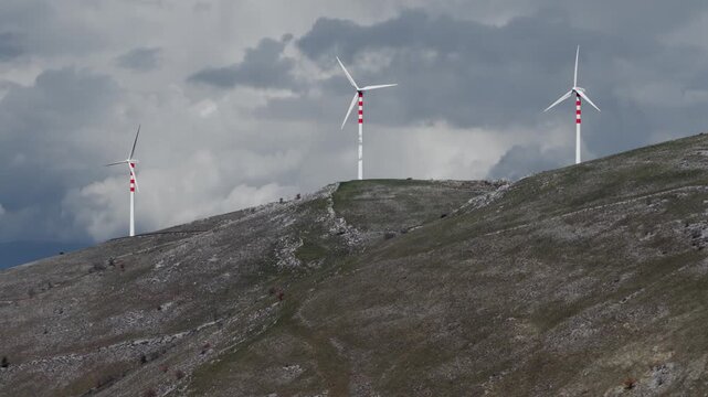 Aerial view of white and red wind turbines standing tall on a grassy hill under a cloudy sky, Cocullo, Abruzzo, Italy.