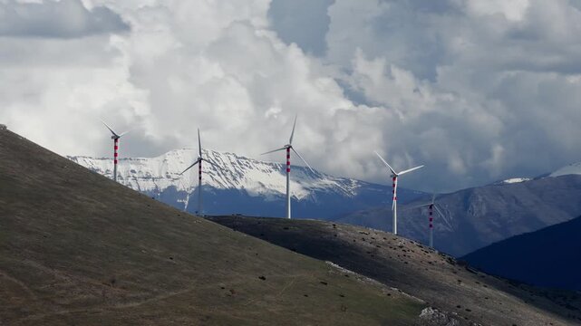 Aerial view of wind turbines standing tall on a grassy hill with snow-capped mountains in the background, Cocullo, Abruzzo, Italy.