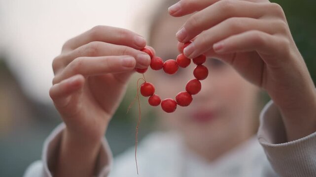 Hands shaping red beads into heart, girl outdoors closeup, delicate fingers forming heart from round beads on thin string soft bokeh background, warm dusk light, nostalgic atmosphere of handmade