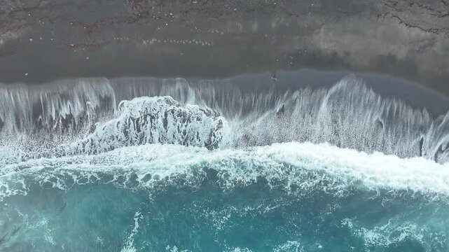 Ocean waves crash onto dark sandy beach, creating foam and ripples, with a clear blue water gradient visible in the foreground and rocky shoreline in the background. Black volcanic sand. Aerial view
