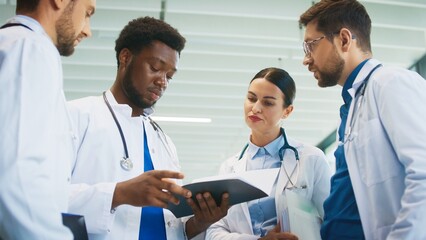 Fototapeta premium Focused male doctor holding chart while colleagues gathering around him in bright hospital hall. Reviewing patient notes. Clarifying duties. Coordinating tasks for busy clinical shift.
