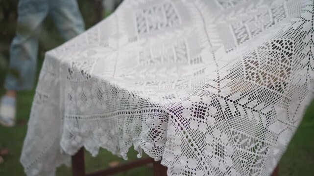 Rustic wooden table draped in lace white woman hostess smoothing delicate crochet cloth over weathered table in garden soft overcast light cozy cottagecore atmosphere hands arranging corners