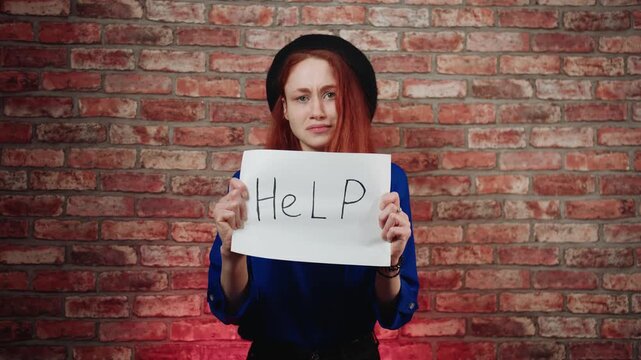 A portrait of a sad caucasian young redhead woman in a blue blouse and black beret, holds a sign on paper "Help" against brick wall. A gesture and request for help to someone in need or a prisoner