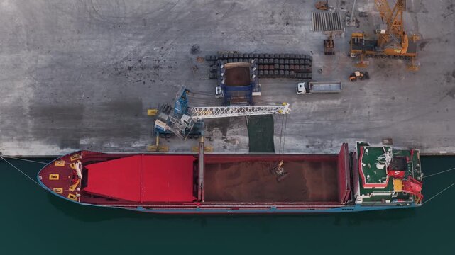 Aerial view of a cargo ship docked in port, its red hull contrasting with the blue-green water, with cranes and cargo nearby, Vasto, Abruzzo, Italy.