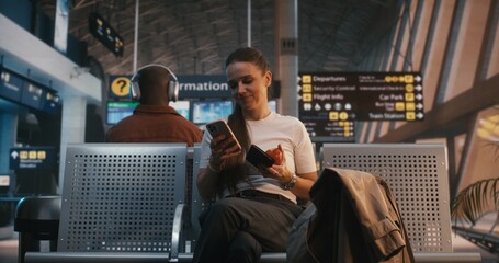 Stylish Female Traveler Sits In Departure Lounge, Holding Boarding Pass And Red Apple. Woman Enjoys Healthy Snack While Scrolling Through Smartphone. Balanced Lifestyle, Modern Travel Habits. © Framestock