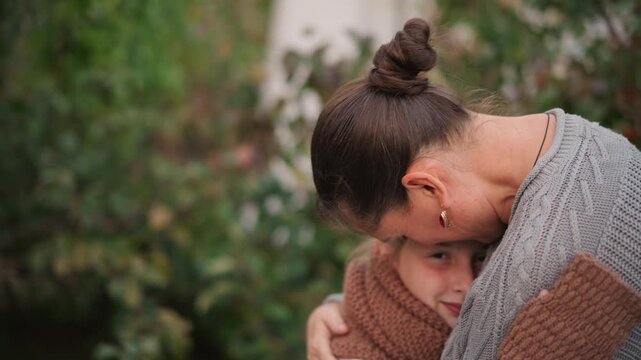 Mother hugging daughter outdoors in garden, cozy knit sweater and scarf, tender forehead kiss, closeup cinematic bokeh, soft warm tones, emotional protective embrace and gentle smile conveying family