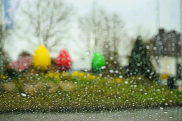 Obraz premium Abstraktes Osterpanorama mit Regentropfenmuster vor Glasscheibe vor bepflanztem Erdhügel mit grünen Narzissen, Gras, Tannen und bunten großen Osterei-Figuren vor Himmel bei Schnee, Sturm im Frühling