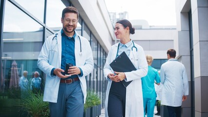 Charming male doctor holding coffee and walking beside smiling female colleague outside clinic....