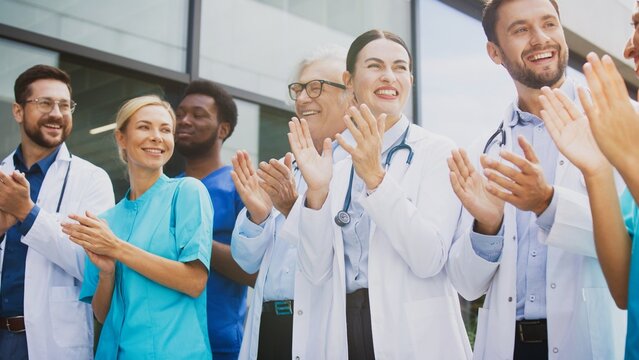Smiling medical staff clapping hands while standing outside clinic under bright daylight. Confident group celebrating progress and sharing uplifting energy. Professionals enjoying supportive moment.
