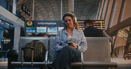 Obraz premium Female Traveler Scans Boarding Pass With Smartphone, Then Puts on Large Over Ear Headphones to Listen Music and Scroll Social Media. Concept of Digital Nomad Lifestyle and Youth Travel Culture.