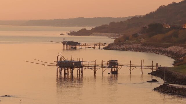 Aerial view of traditional trabocchi fishing platforms stand in the tranquil waters, casting long shadows in San Vito Chietino, Abruzzo, Italy.