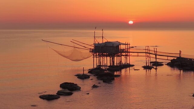 Aerial view of the Trabucco, a traditional fishing structure, stands silhouetted against a golden sunset, San Vito Chietino, Abruzzo, Italy.
