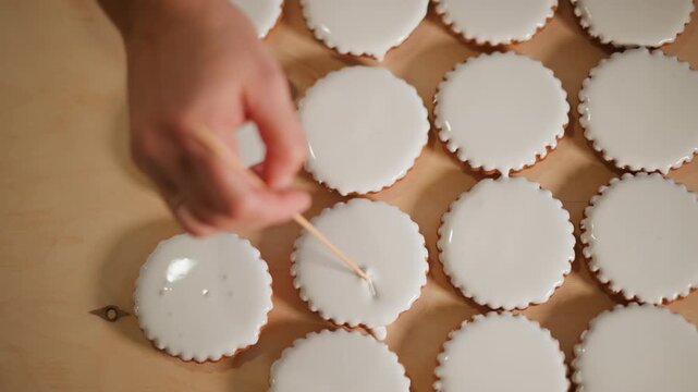 Hand decorating iced mini pies using toothpick, creating delicate pull patterns on smooth white glaze across rows of tart shells on wooden tray, cozy natural light, home bakery closeup