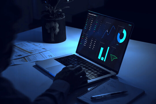 Man working late night at desk analyzing financial trading charts on laptop screen with energy utility bills for domestic budget economy investment concept