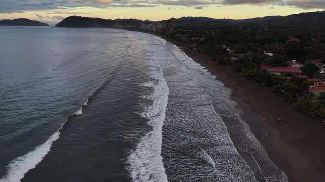 Aerial view of the dark sandy beach meeting the ocean's gentle waves near lush, green vegetation and scattered buildings, Jaco, Costa Rica.