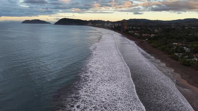 Aerial view of the dark waters meeting the sandy beach, framed by lush greenery and buildings under a pastel sky, Jaco, Costa Rica.