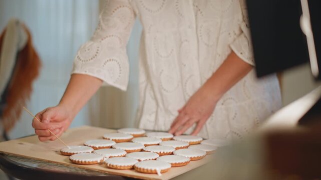 Hands decorating pumpkin cookies on tray, closeup of female baker piping white icing, neat patterns across parchmentlined cookies, warm lamp glow, focused slow motions creating festive seasonal treats