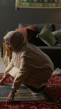 Vertical shot of young Muslim woman wearing light pink hijab while finishing daily prayer of Salah and standing up in modern cozy home with traditional decor