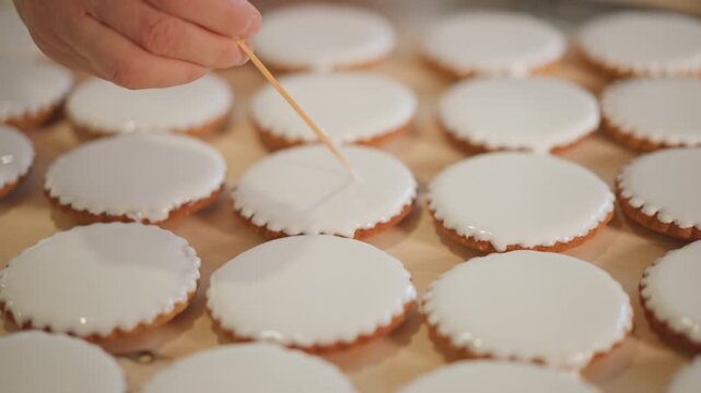 Rows iced mini pies being decorated by quick skilled hands, repetitive pattern across wooden trays, highvolume bakery setting, rhythmic motion, careful dotting of glaze, warm neutral lighting