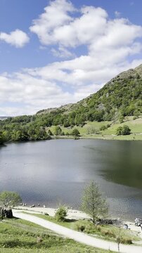 Landscape video of Rydal Water with visible footpath, rolling green hills and trees beneath a partly cloudy blue sky, Lake District, England, UK.
