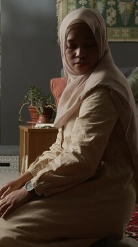 Vertical side view shot of Muslim young woman wearing hijab finishing daily prayer ritual at home turning head during Salah in calm home setting