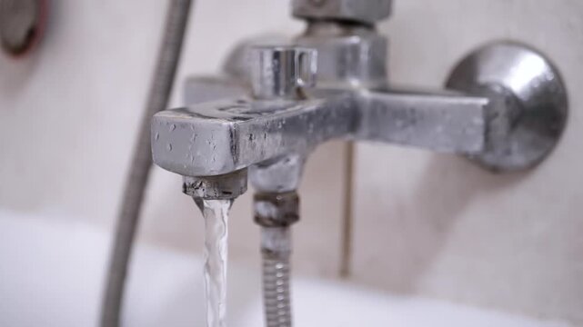 A dirty, rusty bathtub faucet runs, pouring water into the tub. The state of the fixture and tiles suggests a need for home improvement.