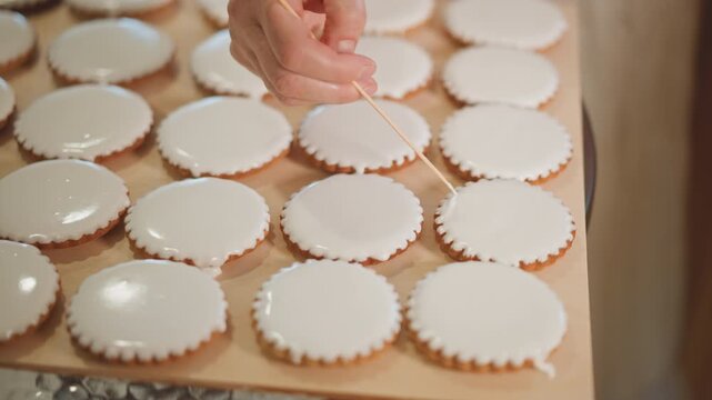 Iced sugar cookies on wooden tray, pastry chef decorating dozens with toothpick. Close view of neat round cookies with smooth white glaze as hand dots and adjusts surface, warm ambient bakery light,