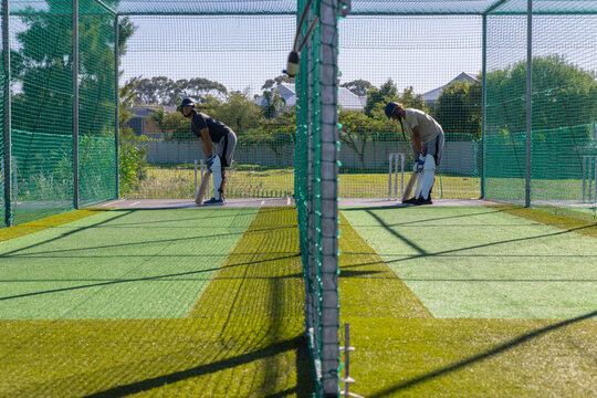 Two men practicing batting in adjacent cricket nets with wooden bats, helmets, pads