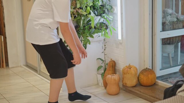 White boy picking up pumpkin by entryway, playful helper carrying squash toward kitchen for pie prep, casual shorts and shirt, sunlight on tile, candid harvest chore and family prep scene