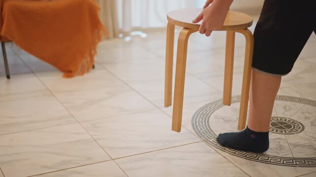 Small wooden stool on tiled floor, person steps forward and picks up stool, patterned medallion tile underfoot, orange throw on sofa, natural morning light, gentle domestic action in simple interior