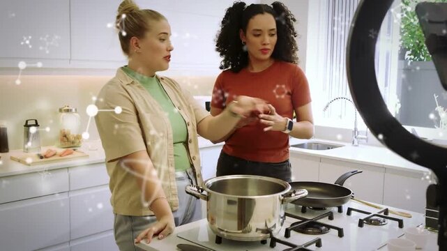 Two cooking hosts reading phone pouring oil, prepping pan with spatula while particles overlaying
