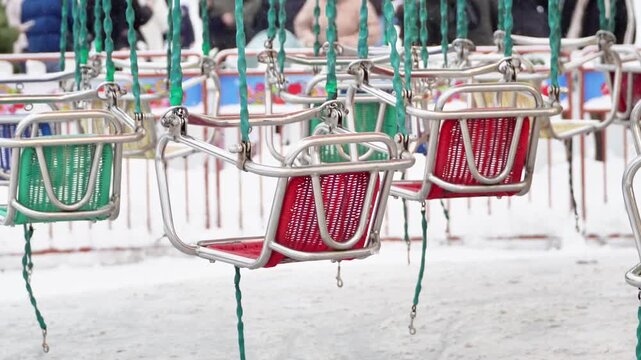 Empty colorful swing seats of a chain carousel ride at an amusement park in winter. Row of vacant colorful chain swing ride seats covered in snow concept of offseason, solitude and closed theme park. 