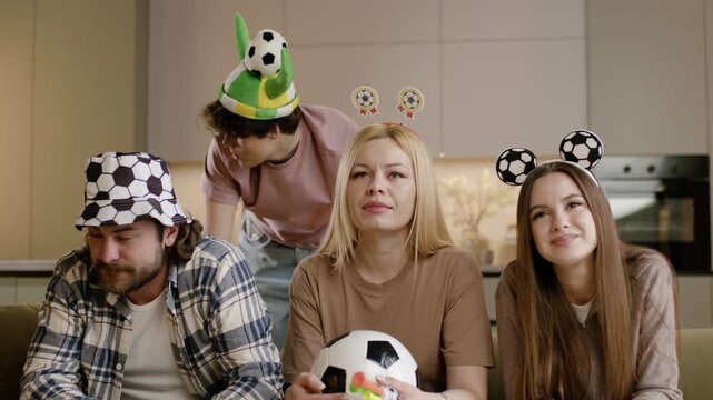 Group of four friends wearing soccer-themed hats and headbands, sitting on a couch, engaged in watching a soccer game with a soccer ball in hand