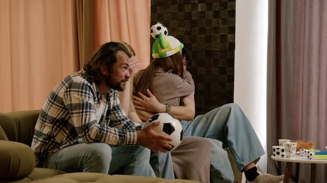 Group of friends celebrating a soccer match at home, showing excitement and joy while holding soccer balls and wearing festive hats in a cozy living room setting