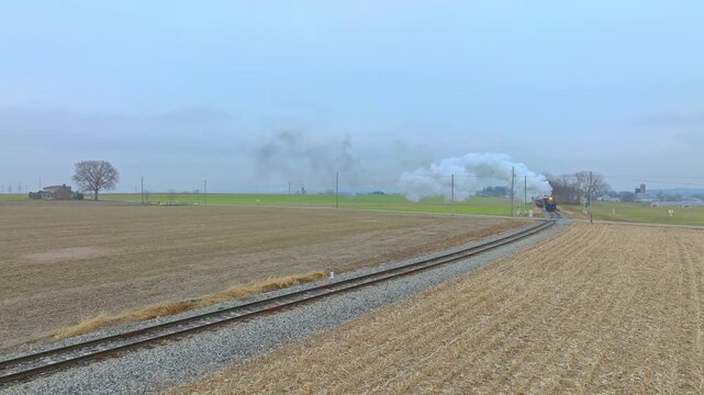 A steam train moves along the tracks in a rural area. The train releases smoke as it travels through open fields. The sky is overcast, and the landscape is mostly flat.