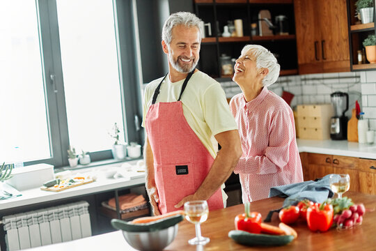 Portrait of happy senior mid aged mature couple prepering meal with fresh vegatebles in kitchen at hpme
