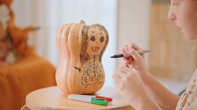 White girl drawing on pumpkin face, indoor cozy scene with wooden stool, markers and crayons on table, focused expression, warm soft light, autumn decor, hands adding smiling face and floral patterns